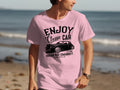 On a sandy beach, someone wears a light pink Garment Graphics t-shirt featuring Enjoy Classic Car and Vintage Ride Community above a classic car illustration—an ideal spot for car lovers amidst soft sand and ocean waves.