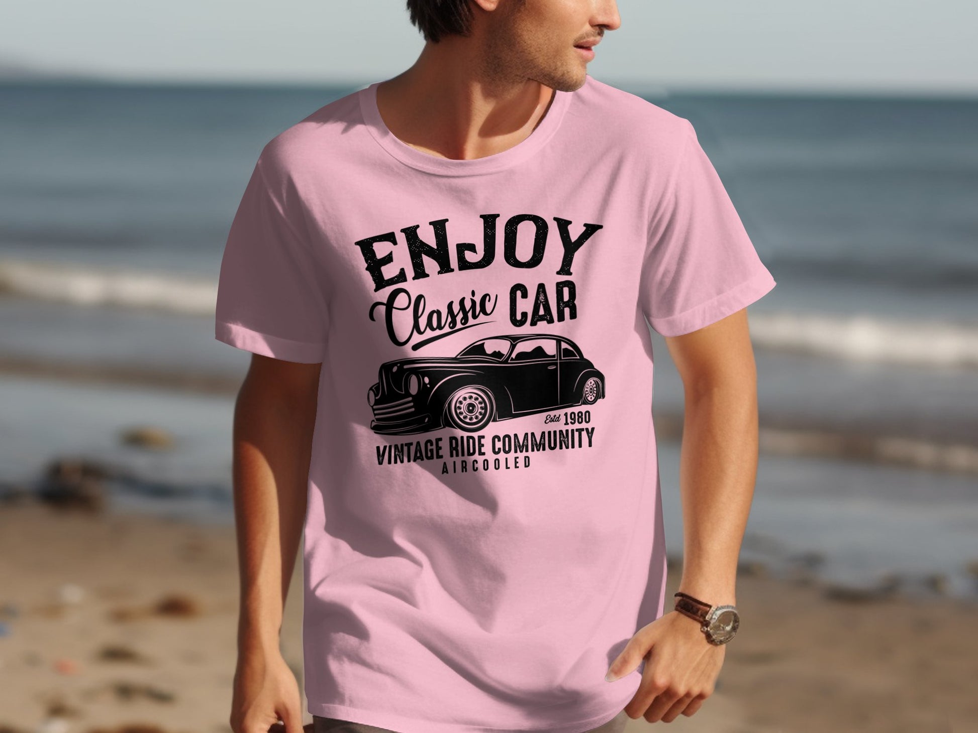 On a sandy beach, someone wears a light pink Garment Graphics t-shirt featuring Enjoy Classic Car and Vintage Ride Community above a classic car illustration—an ideal spot for car lovers amidst soft sand and ocean waves.