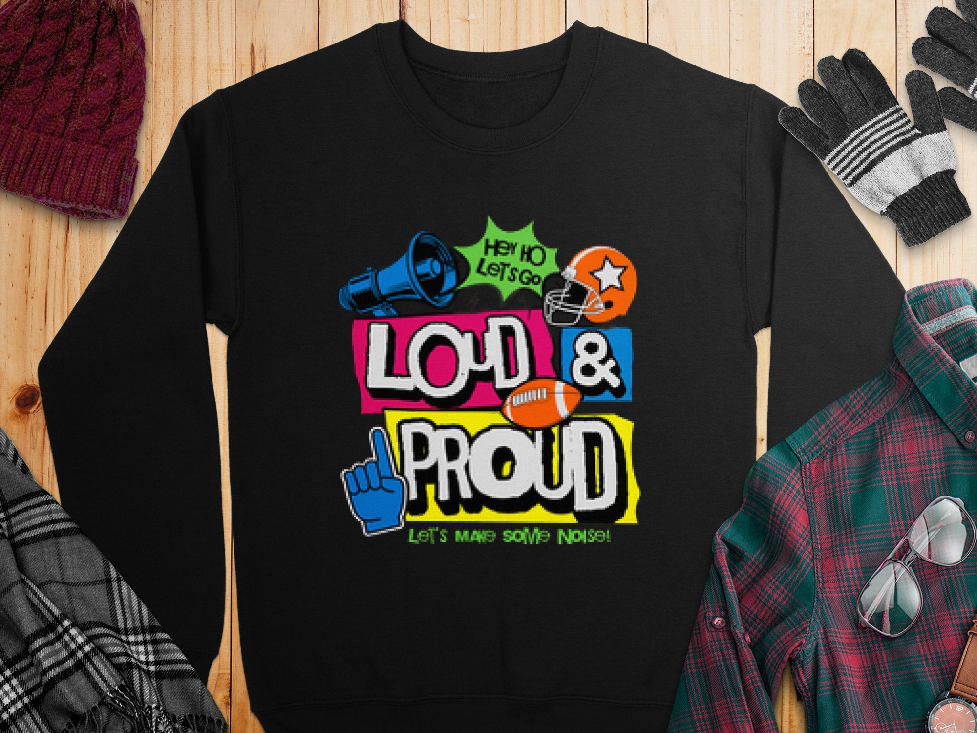 A unisex black sweatshirt from Garment Graphics features the vibrant Hey Ho Lets Go Loud & Proud design, showcasing megaphone, foam finger, football, and helmet graphics. Its part of the unique style of Loud and Proud Sweatshirts, set against a wooden backdrop with a hat and gloves.