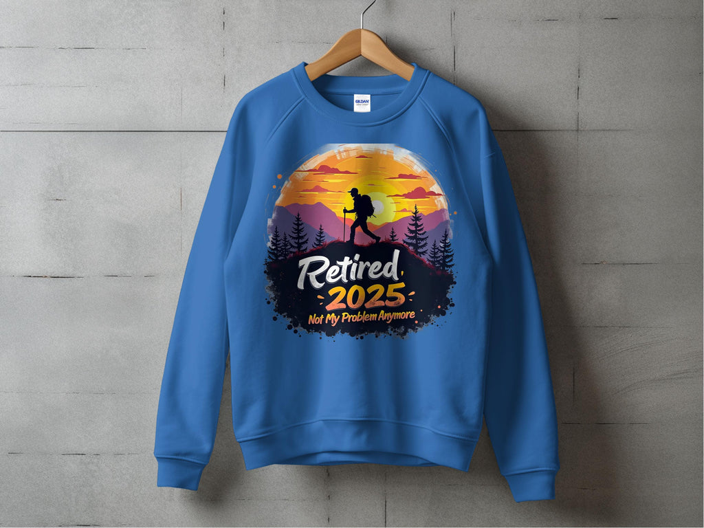 A blue Garment Graphics sweatshirt hangs on a wooden hanger against a gray wall, featuring a hiker silhouette framed by a sunset and mountains. The bold, decorative text reads: Retired 2025: Not My Problem Anymore.