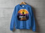 A blue Garment Graphics sweatshirt hangs on a wooden hanger against a gray wall, featuring a hiker silhouette framed by a sunset and mountains. The bold, decorative text reads: Retired 2025: Not My Problem Anymore.