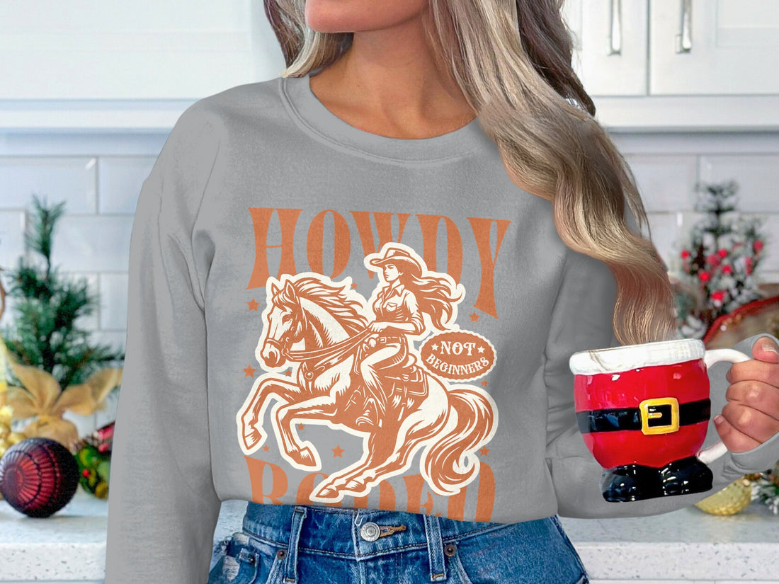 A person outdoors wears a white Garment Graphics sweatshirt with a brown cowgirl and horse graphic, surrounded by stars and featuring the words Howdy Rodeo and Not for Beginners. Perfect for rodeo enthusiasts looking to embrace the cowboy spirit in style.