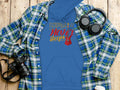 A classic fit blue hoodie by Garment Graphics, featuring Music Never Sleeps and a guitar design, is displayed on a wooden surface. Its surrounded by a blue-green plaid shirt, headphones, a camera, coffee cup, and notebook—ideal for music hoodie enthusiasts!.