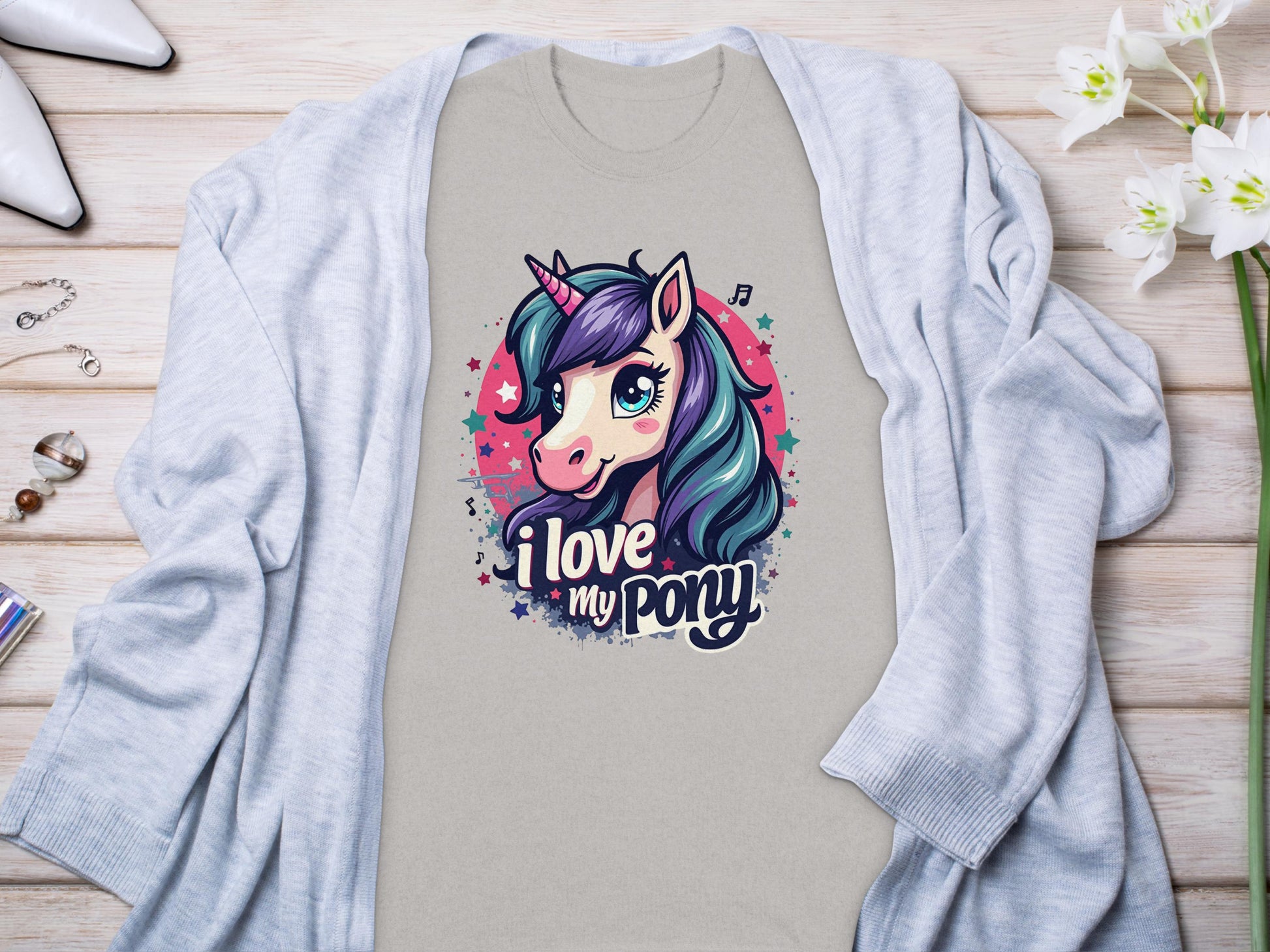 A beige pony graphic T-shirt by Garment Graphics features a vibrant unicorn with the phrase I love my pony. Paired under a light gray cardigan on light wood, its beautifully accented with flowers and small accessories, ideal for any unicorn enthusiast.