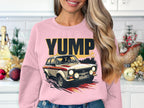 A woman with long hair dons a pink Garment Graphics sweatshirt featuring a YUMP rally car design in her kitchen, where holiday decorations scattered on the counter encapsulate the festive spirit.