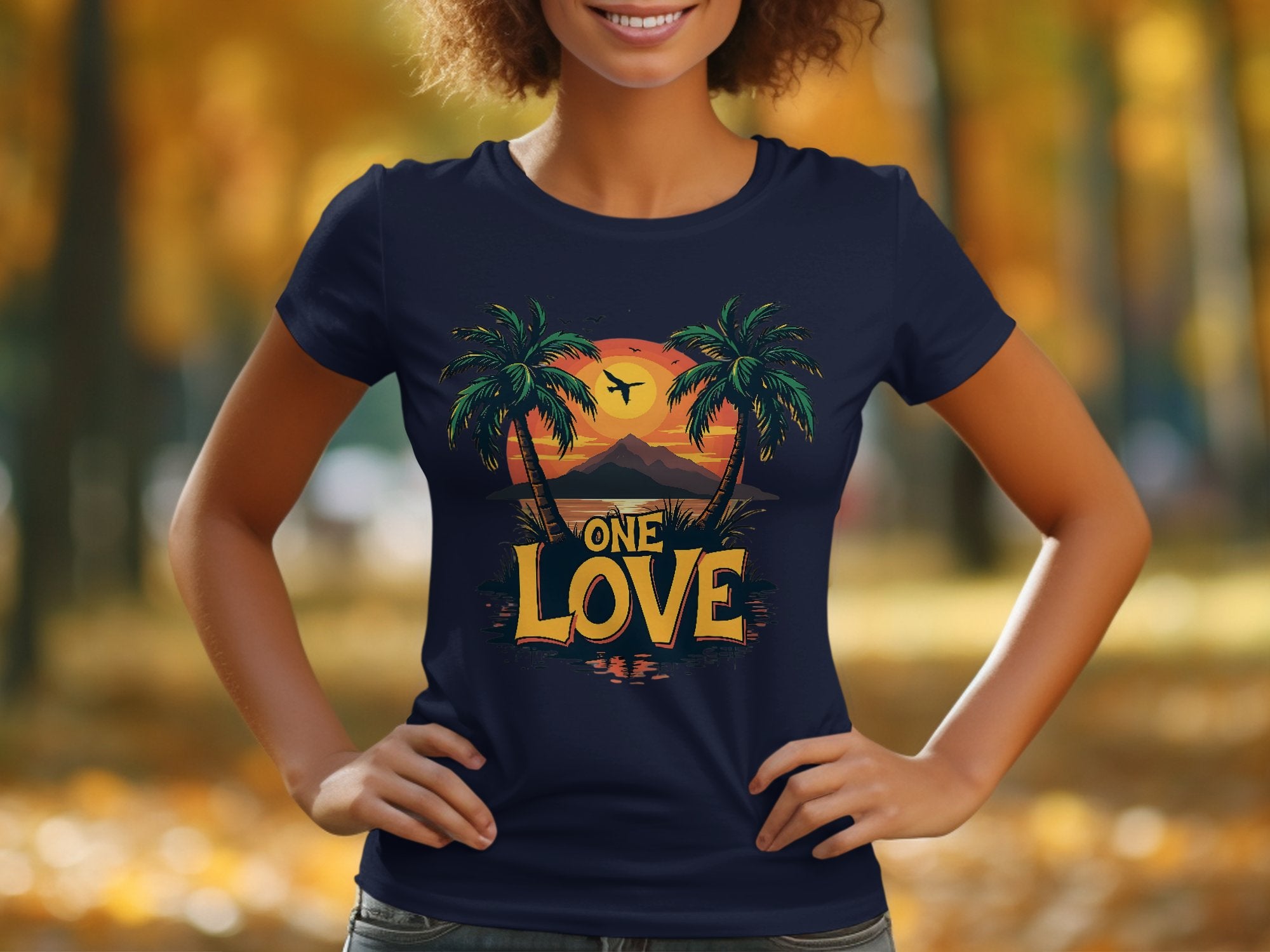 In a park-like setting, someone wears a Garment Graphics navy T-shirt featuring two palm trees, an airplane, and a tropical sunset. The words One Love evoke a carefree beach outing vibe amidst the backdrop of blurred autumnal trees glowing with warm yellow and orange leaves.