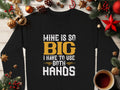 A black Garment Graphics shirt boldly displays Mine Is So Big I Have To Use Both Hands in white and yellow, set on a wooden surface with Christmas decorations and a steaming coffee, capturing festive essence.
