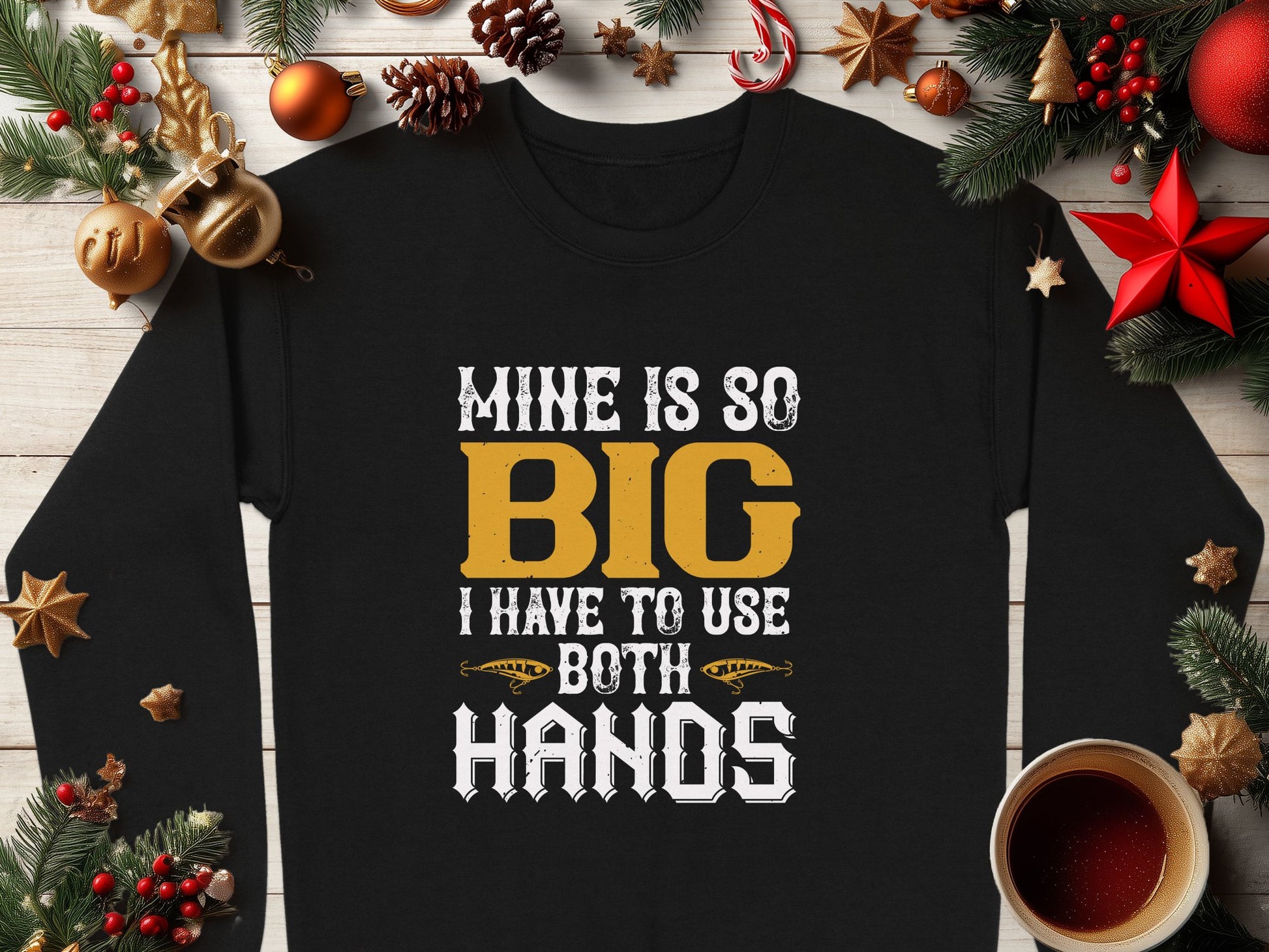 A black Garment Graphics shirt boldly displays Mine Is So Big I Have To Use Both Hands in white and yellow, set on a wooden surface with Christmas decorations and a steaming coffee, capturing festive essence.