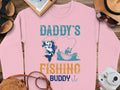 A pink Garment Graphics sweatshirt with Daddys Fishing Buddy and a boat fishing graphic is perfect for fishing enthusiasts. Displayed among a camera, sandals, notebook, straw hat, and sunglasses on a wooden surface, its ideal for fishing adventure lovers.