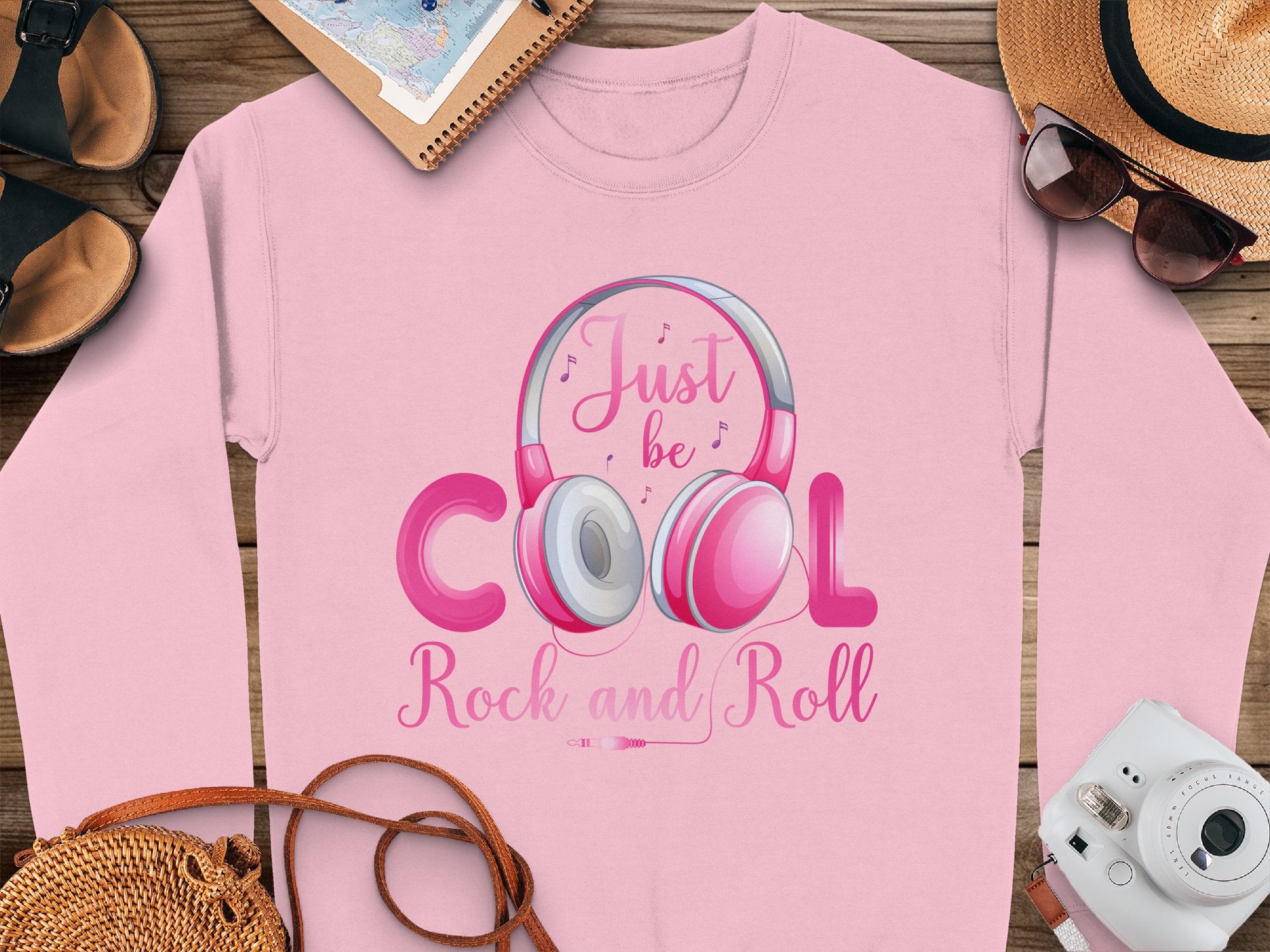 A Garment Graphics pink sweatshirt sits on a wooden surface, showcasing Rock and Roll Headphones with the Just be Cool Rock and Roll slogan. Perfect for music lovers, its accompanied by a straw hat, sunglasses, notebook, sandals, and a camera.
