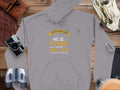 Garment Graphics gray fishing hoodie showcases Father and Son Fishing Partners for Life in yellow and white, along with fishing imagery. Featured with a vintage camera, binoculars, coiled rope, sunglasses, and a leather pouch on wood—a must-have for fishing enthusiasts.