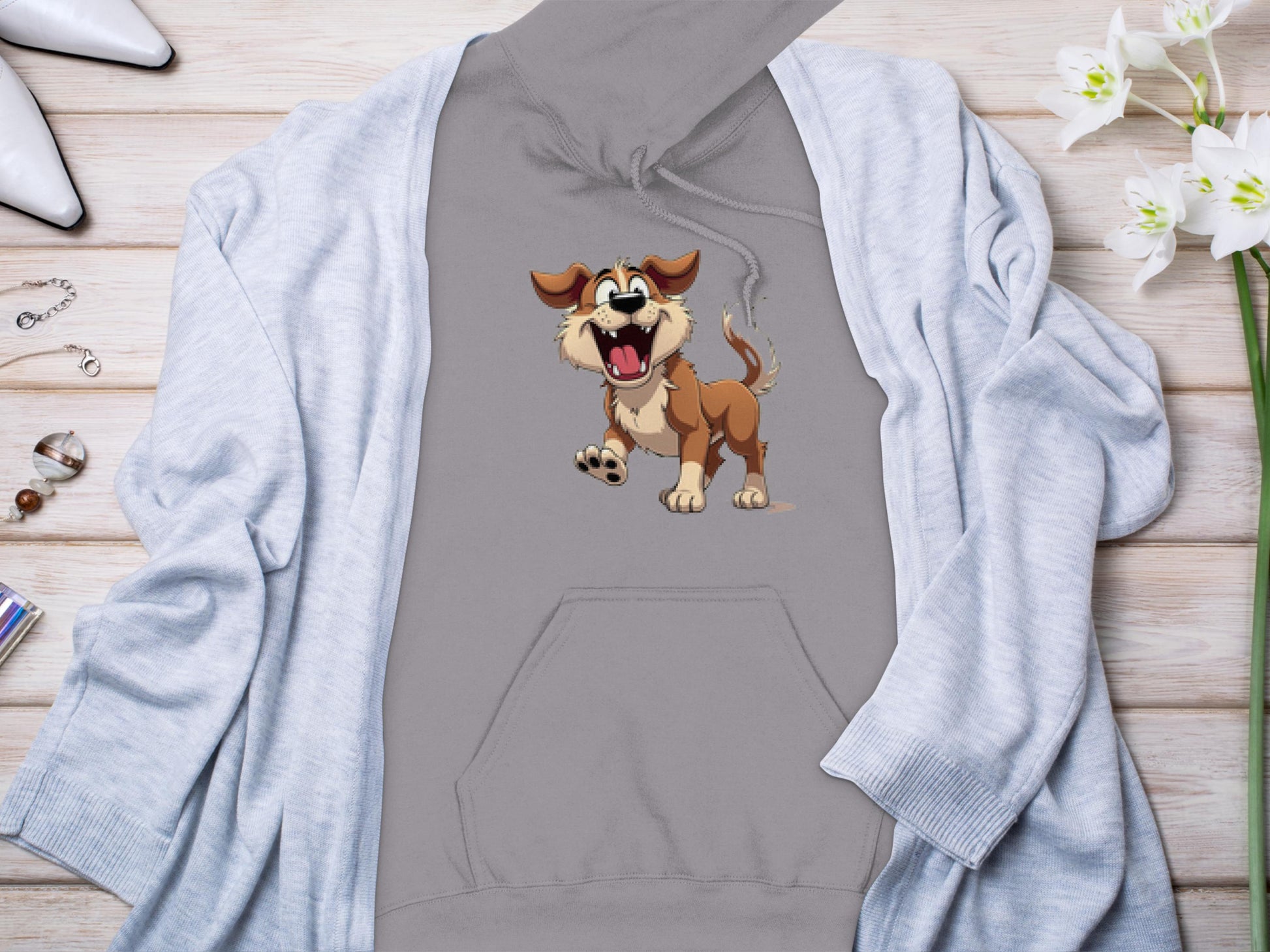 This playful Garment Graphics gray hoodie, perfect for dog lovers with its cartoon dog design, is displayed on a wooden surface. Its attractively complemented by a light gray cardigan, white pointed shoes, jewelry, and a bouquet of white flowers.