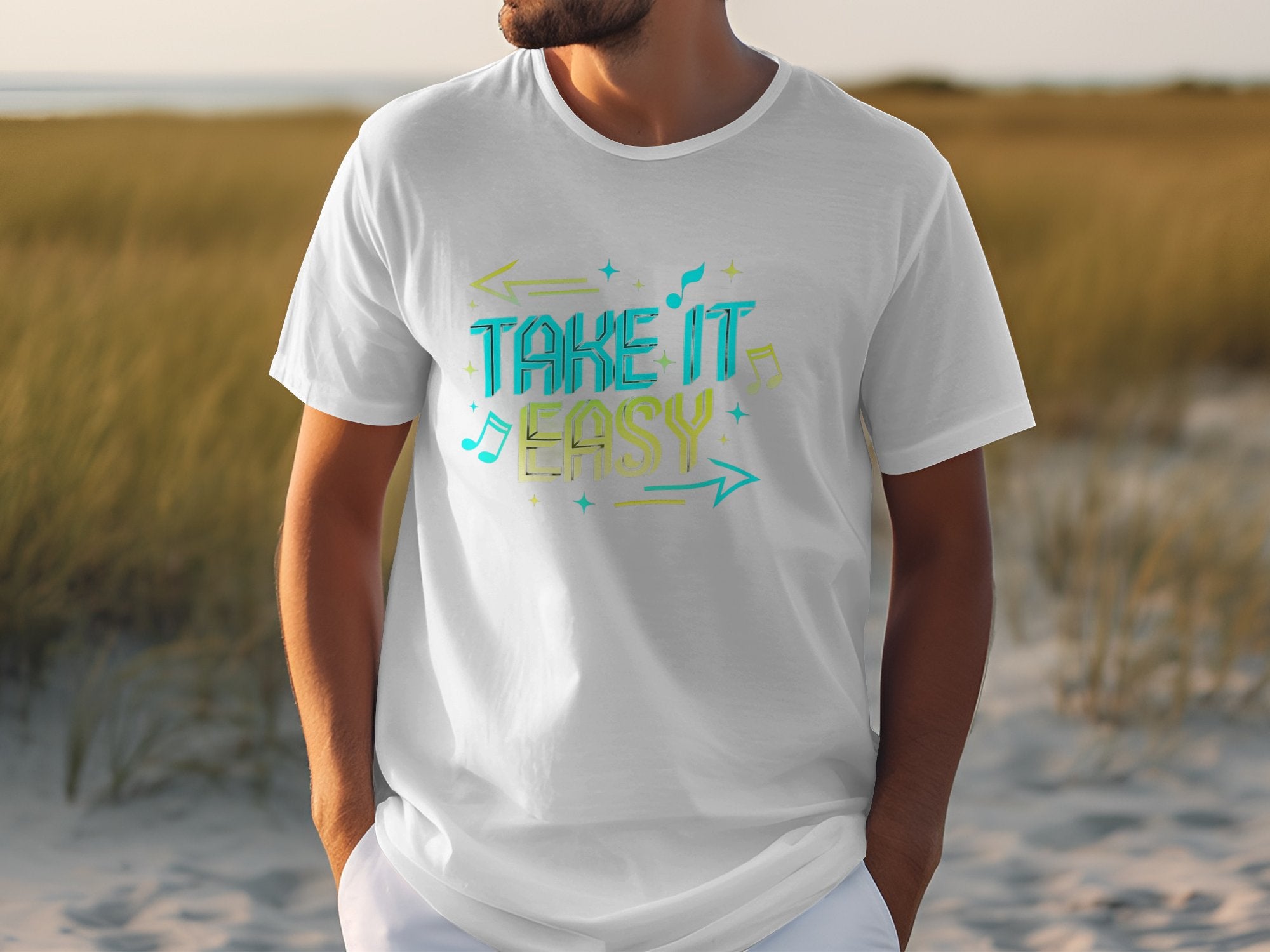 A person in a white Garment Graphics TAKE IT EASY T-shirt, featuring colorful designs and musical notes, stands on a sandy beach with grassy patches leading to the sea.