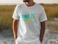 A person in a white Garment Graphics TAKE IT EASY T-shirt, featuring colorful designs and musical notes, stands on a sandy beach with grassy patches leading to the sea.