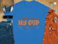 A Garment Graphics blue graphic T-shirt with Youre My Cup of Tea in orange letters sits on a wooden surface, accompanied by headphones, a navy cap, sunglasses, jeans, an orange button-up shirt, a watch, and keys.