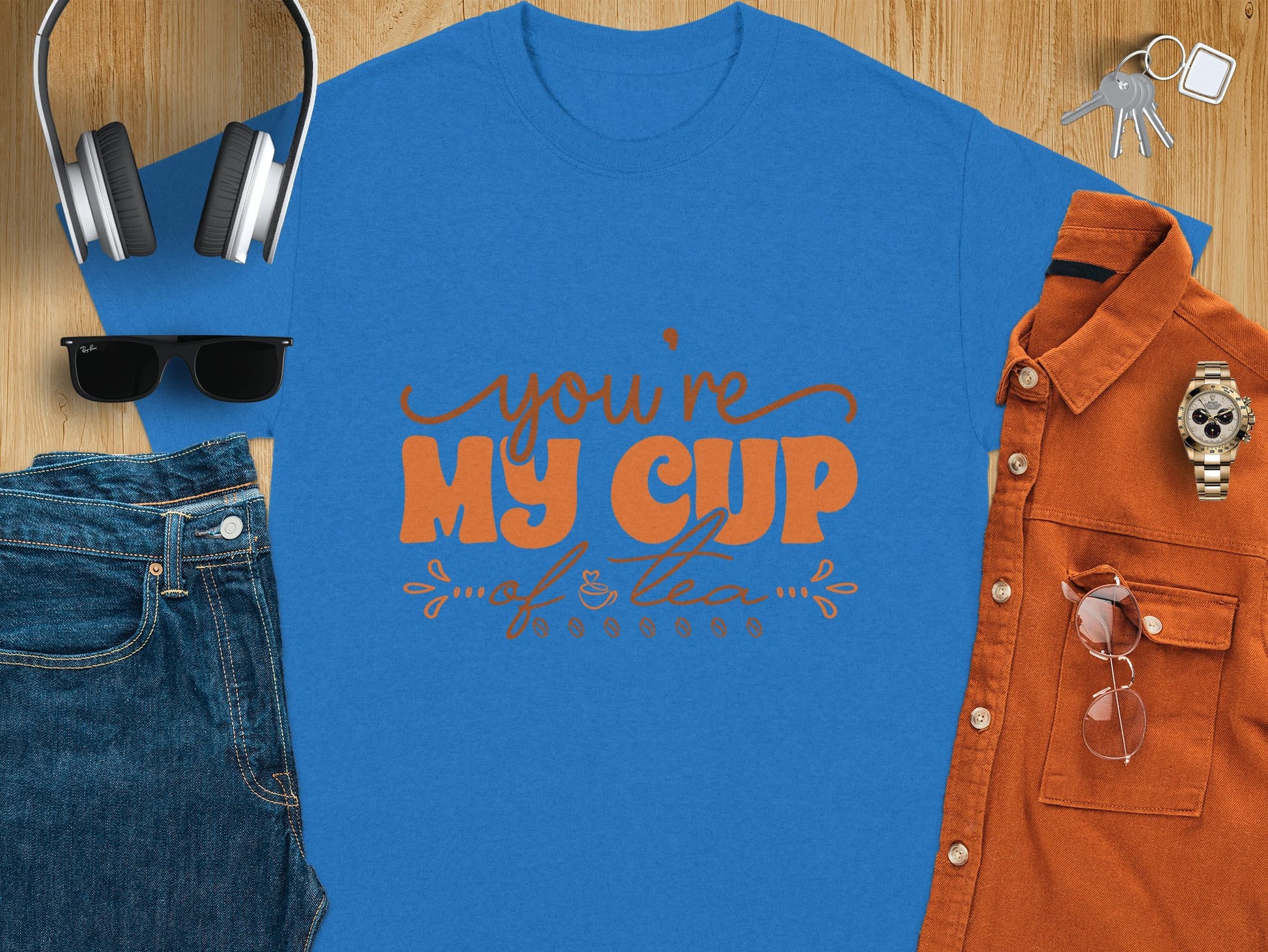 A Garment Graphics blue graphic T-shirt with Youre My Cup of Tea in orange letters sits on a wooden surface, accompanied by headphones, a navy cap, sunglasses, jeans, an orange button-up shirt, a watch, and keys.