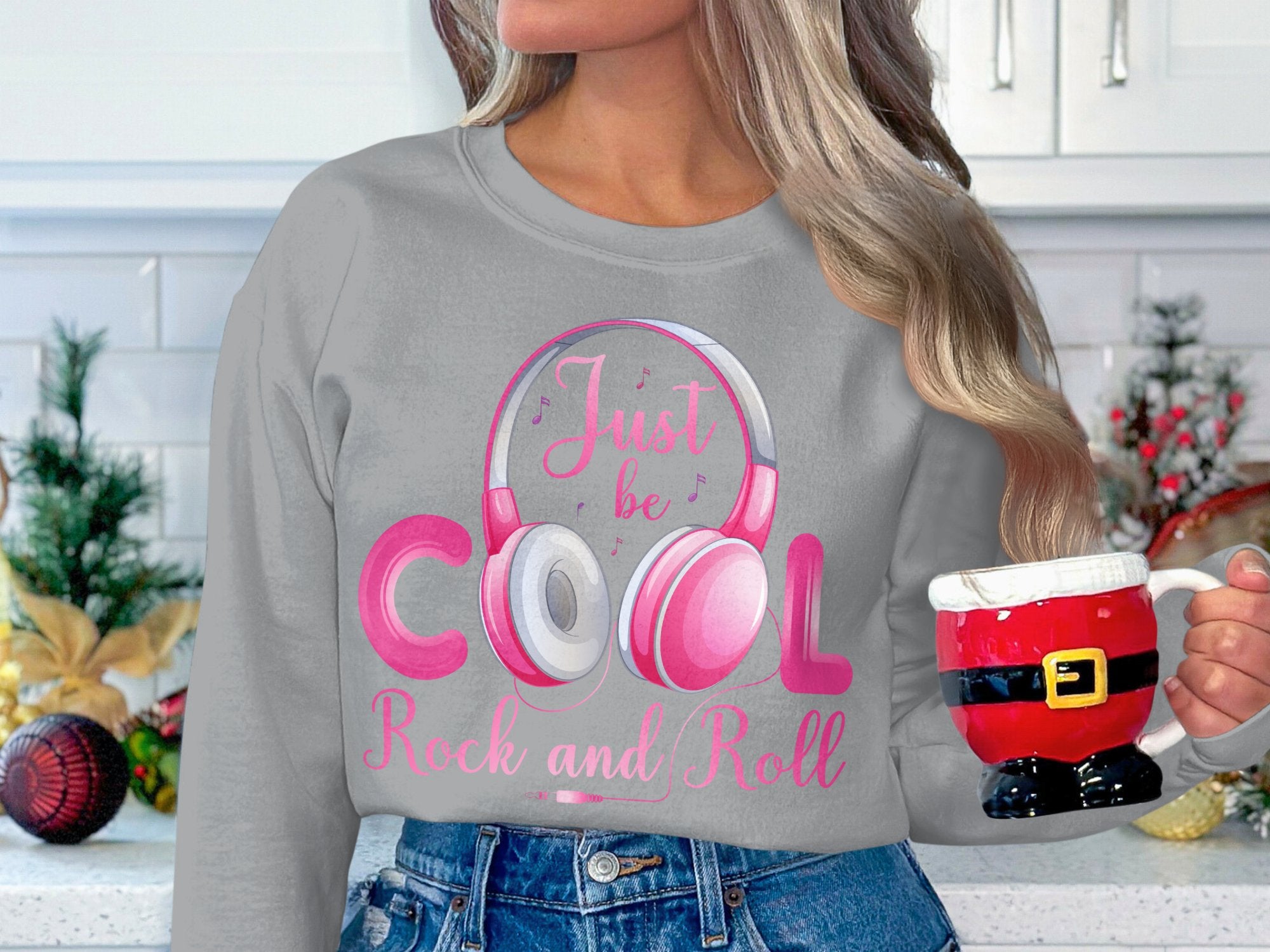In a place filled with festive cheer, someone holding a Santa-themed mug radiates relaxed vibes perfect for music fans. They wear pink headphones and a gray Just be Cool Rock and Roll sweatshirt from Garment Graphics, surrounded by holiday decor and plants, creating a stylish setting.