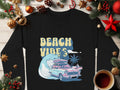 Garment Graphics black sweatshirt showcases a pastel classic car with surfboards over ocean waves, topped by Beach Vibes. The retro scene is festively adorned with pine branches, ornaments, and a coffee cup.