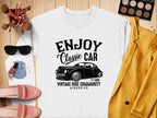 A Garment Graphics white T-shirt with a black classic car graphic and Enjoy Classic Car Vintage Ride Community Aircooled Estd. 1980 rests on wood, surrounded by sunglasses, makeup, a yellow jacket, pink shoes, and brushes for a retro vibe.