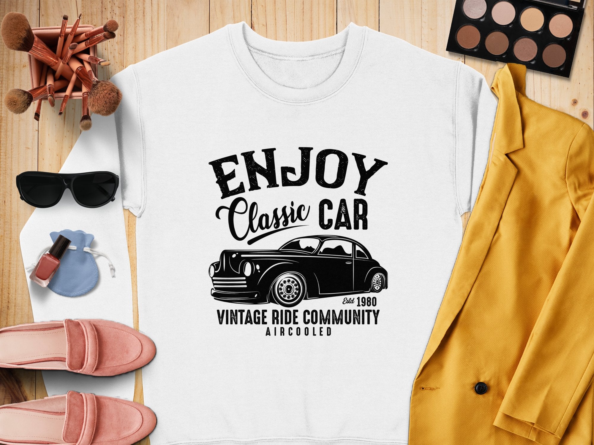 A Garment Graphics white T-shirt with a black classic car graphic and Enjoy Classic Car Vintage Ride Community Aircooled Estd. 1980 rests on wood, surrounded by sunglasses, makeup, a yellow jacket, pink shoes, and brushes for a retro vibe.