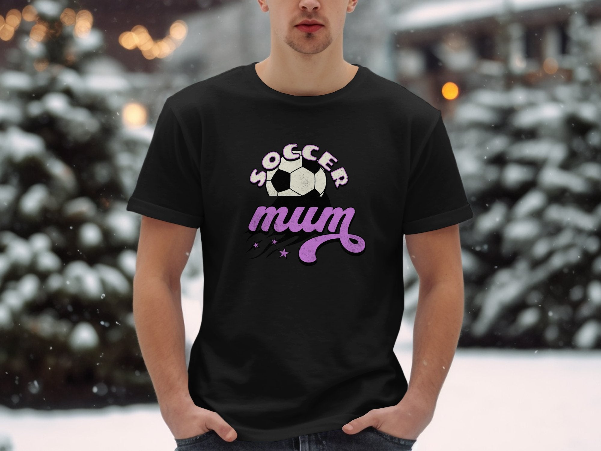 In a snowy outdoor scene, someone wears a Garment Graphics classic-fit black T-shirt with Soccer mum and a soccer ball graphic. Trees and snow create a scenic background.