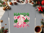 The Garment Graphics Wild Side Sweatshirt displays bold graphics of a person in a cowboy hat, finger to lips, surrounded by flowers with HANDS OFF! in green and pink. Pictured with Christmas ornaments and coffee, this sweatshirt offers unisex sizing to fit everyone.
