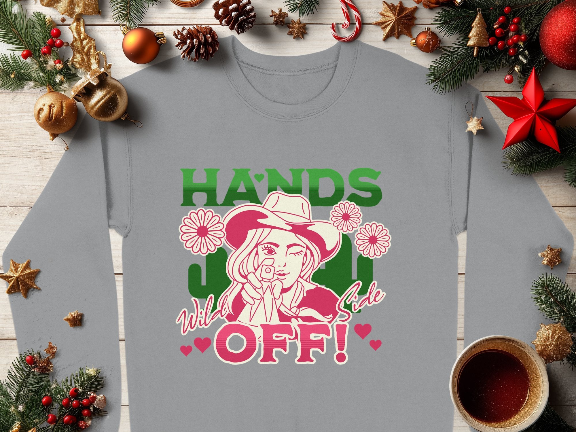 The Garment Graphics Wild Side Sweatshirt displays bold graphics of a person in a cowboy hat, finger to lips, surrounded by flowers with HANDS OFF! in green and pink. Pictured with Christmas ornaments and coffee, this sweatshirt offers unisex sizing to fit everyone.