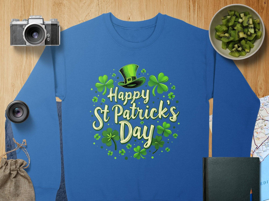 Garment Graphics blue shamrock graphic sweatshirt showcases Happy St Patricks Day text with green shamrocks and a top hat. A camera, lens, cheerful sack, kiwifruit bowl, closed book, and map on the wooden surface complete the vibrant scene.