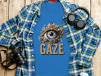 A classic fit Garment Graphics T-shirt with an eye gaze design and GAZE text lies on wood. Its under a blue plaid shirt, surrounded by a camera, headphones, and a white paper cup.