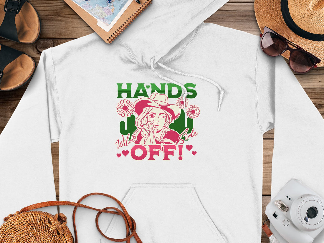 A person wears a Garment Graphics pink Graphic Hoodie with a woman in a hat, HANDS OFF! text in bold green and red, featuring flowers and hearts. Seated, the person pairs it with checkered pants.