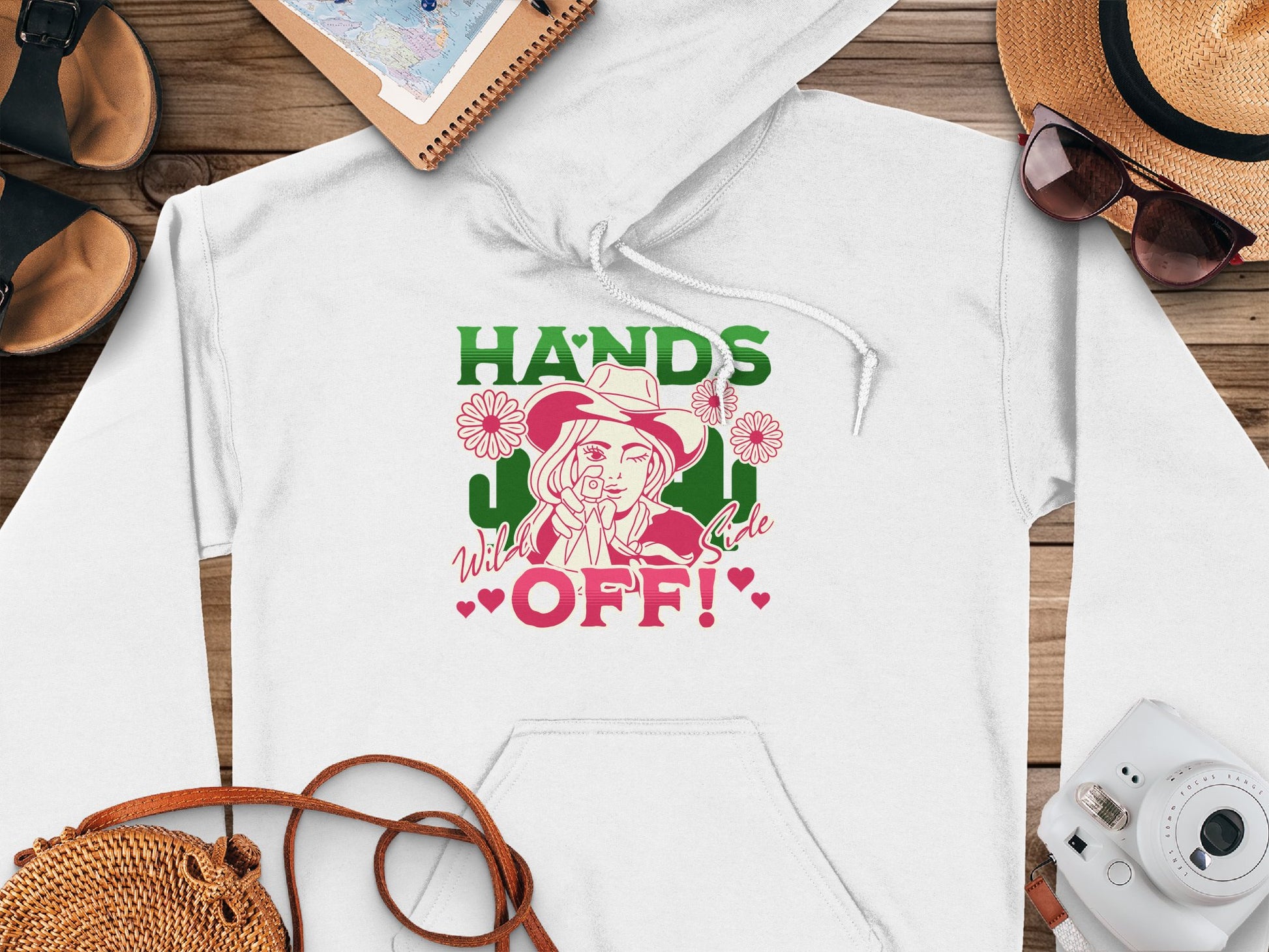 The Garment Graphics hoodie features a bold HANDS OFF! design in green and red. A woman in a hat holding a flower is prominently displayed on this white graphic hoodie, styled with accessories like a hat, camera, sandals, and notebook on a wooden surface.