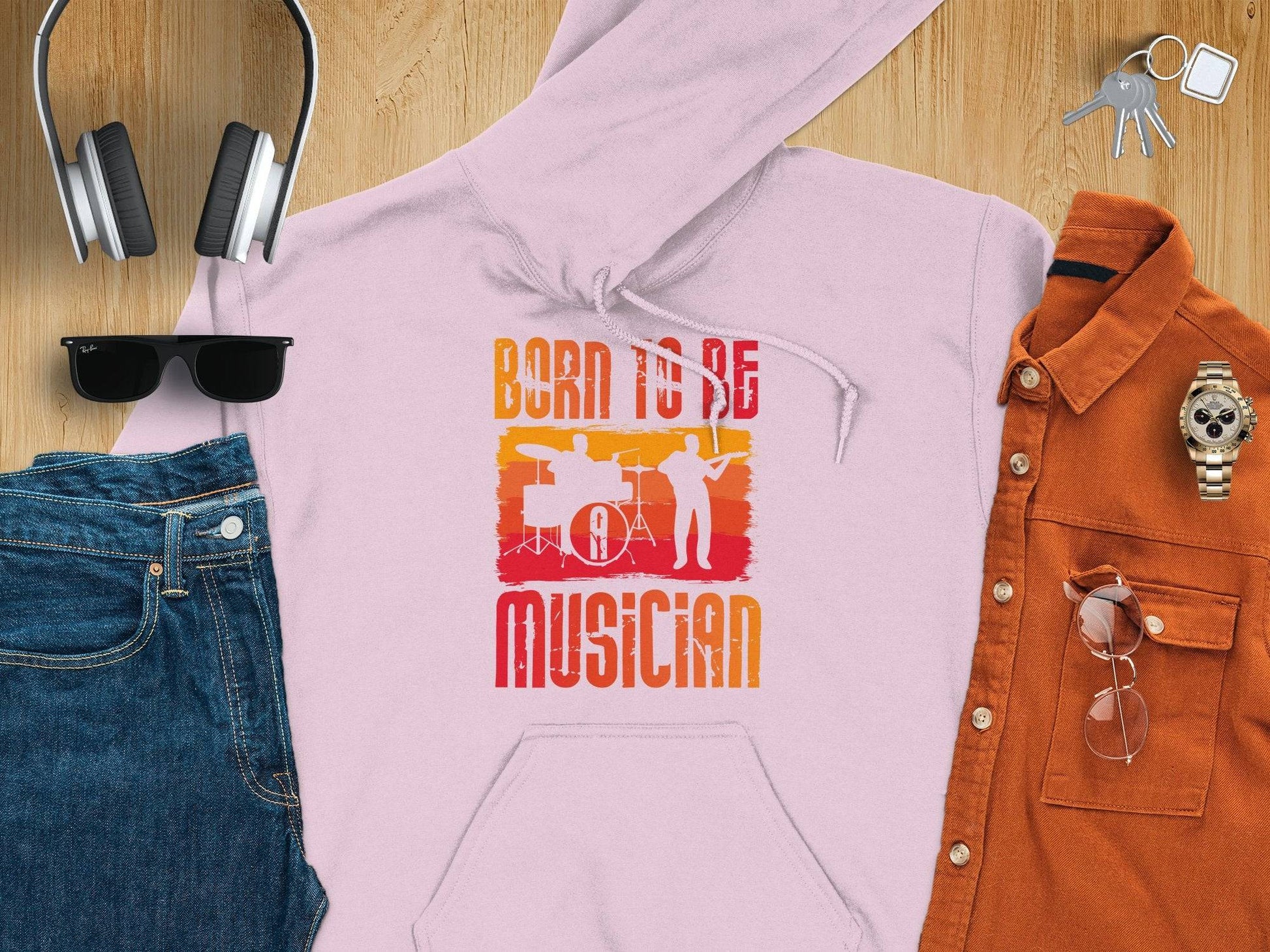 A light pink unisex fashion hoodie by Garment Graphics features a Born to Be Musician design with silhouettes of two musicians. Ideal for music lovers, it pairs perfectly with folded jeans, sunglasses, headphones, keys, a watch, and an orange button-up shirt on a wooden surface.