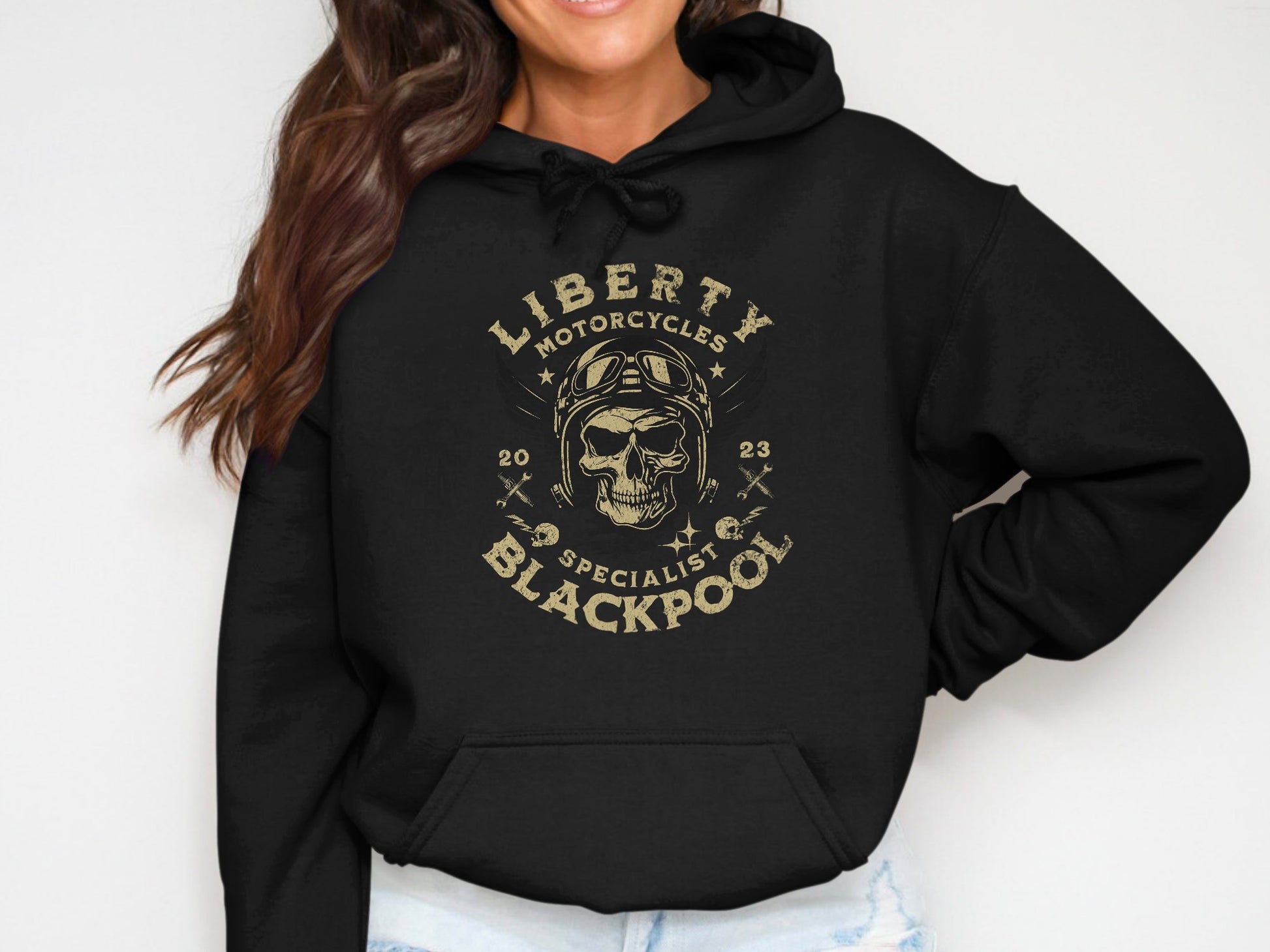 A woman wears a Garment Graphics Blackpool Hoodie with a unique skull, helmet, and wrenches design, featuring the text Liberty Motorcycles Specialist Blackpool 2023, embodying the brands distinctive style.