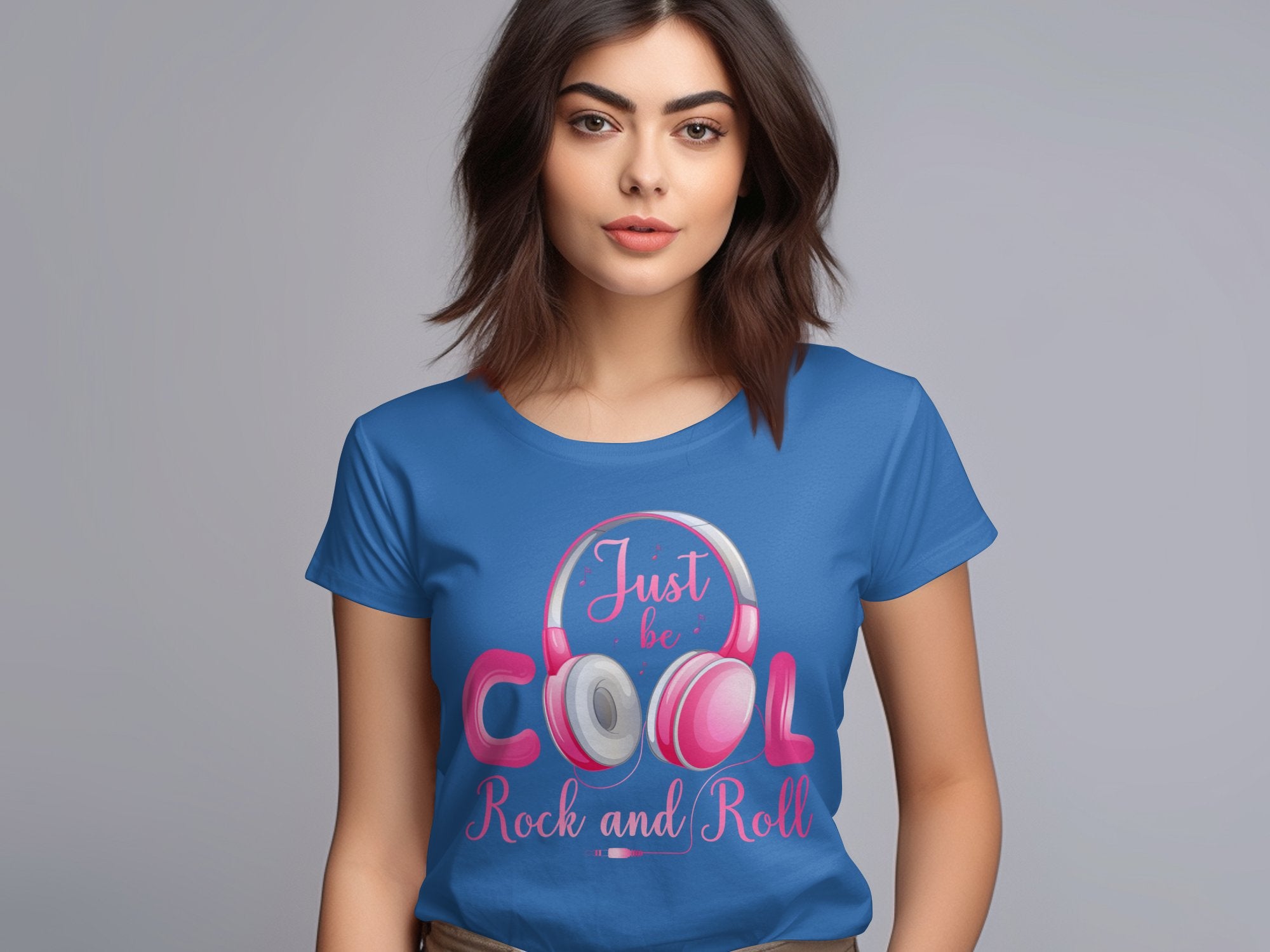 A woman with shoulder-length brown hair stands against gray, wearing a blue t-shirt from Garment Graphics stating Just be COOL Rock and Roll. Featuring white and pink headphones, its perfect for music lovers who appreciate headphone-themed tees.