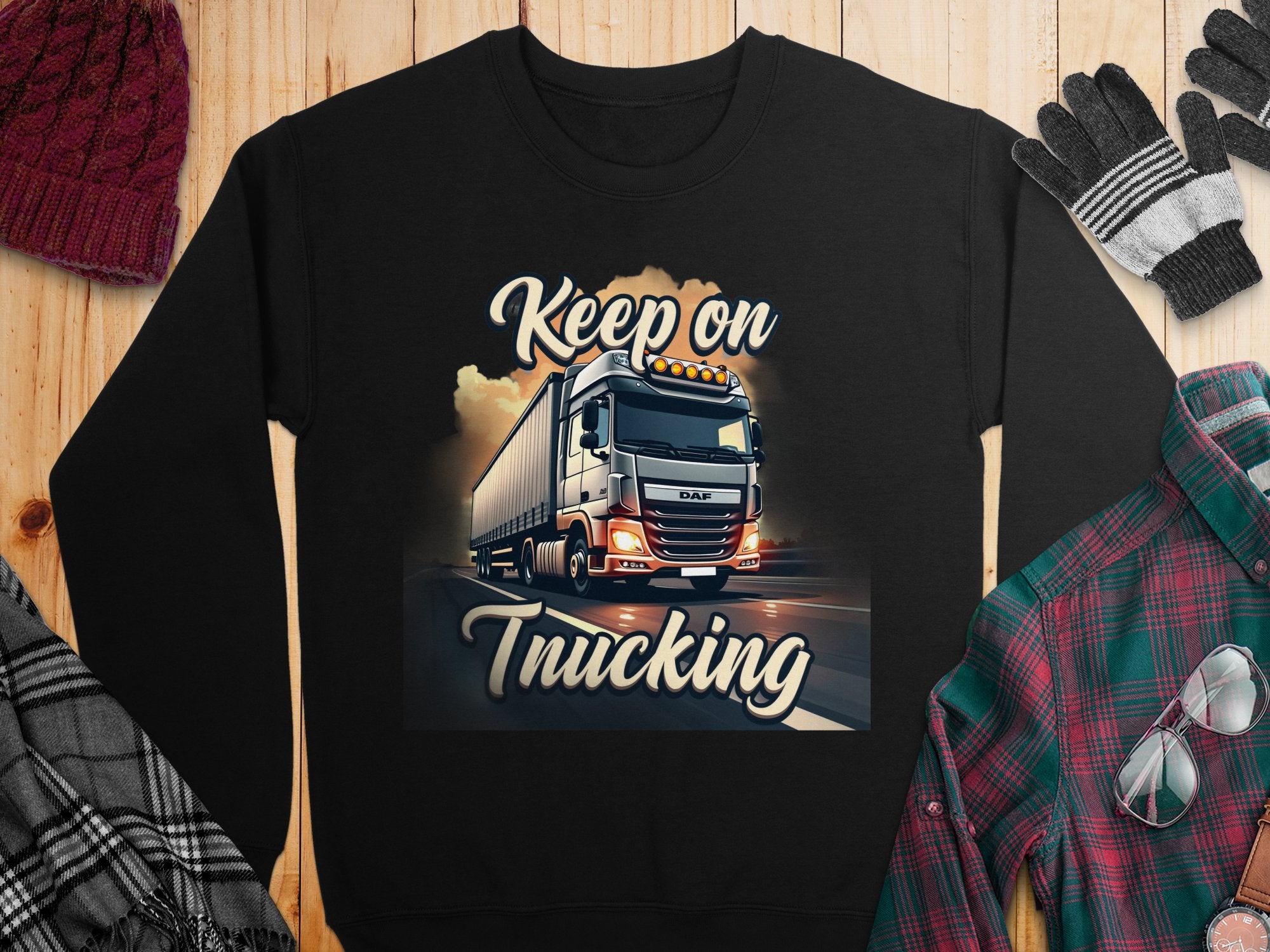 A black Garment Graphics sweatshirt with a truck design and Keep on Trucking text is displayed on wood. Unisex sizing, its styled with a plaid shirt, gloves, watch, and knitted hat for a rugged look.