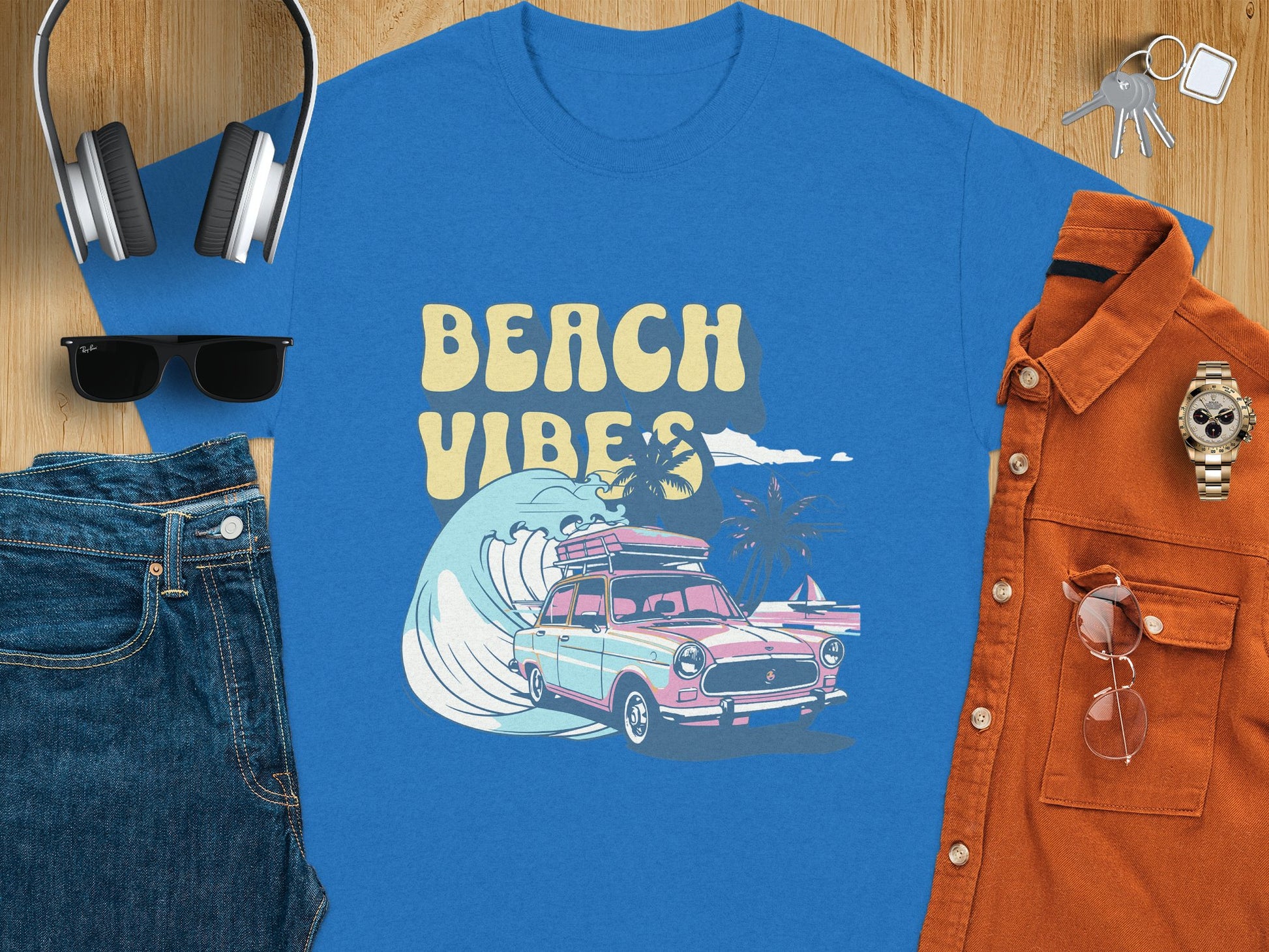 A Garment Graphics t-shirt featuring a Beach Vibes design with a classic car riding a wave is paired with essentials like headphones, keys, sunglasses, and jeans on a wooden surface for the ultimate laid-back look.