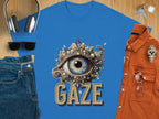 A classic blue T-shirt by Garment Graphics, with an ornate eye gaze design adorned with pearls and jewels above GAZE. Its paired with headphones, sunglasses, jeans, a watch, rust-colored shirt, keys, and a smartphone on a wooden surface.