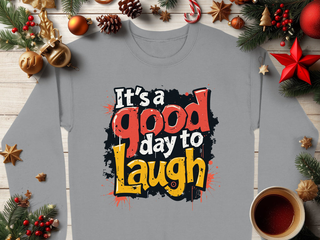 A Garment Graphics gray Graphic Art Sweatshirt, featuring Its a Good Day to Laugh in bold letters. Styled with Christmas decorations like pine branches, ornaments, and a hot beverage cup on wood for coziness. Unisex sizing.