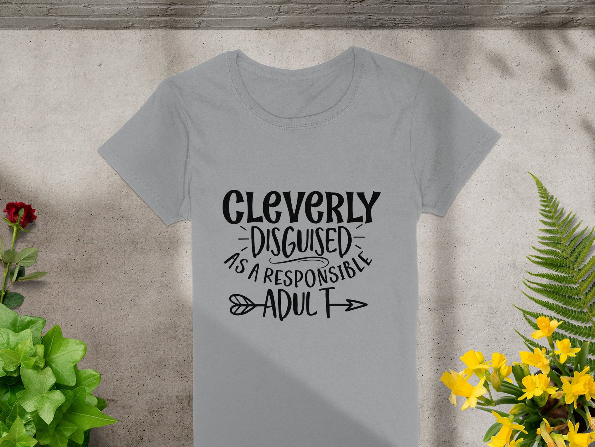 A Garment Graphics light gray, semi-fitted T-shirt lies on concrete, boldly featuring Cleverly Disguised as a Responsible Adult in black text. Green plants and yellow flowers frame the image, adding a natural flair.