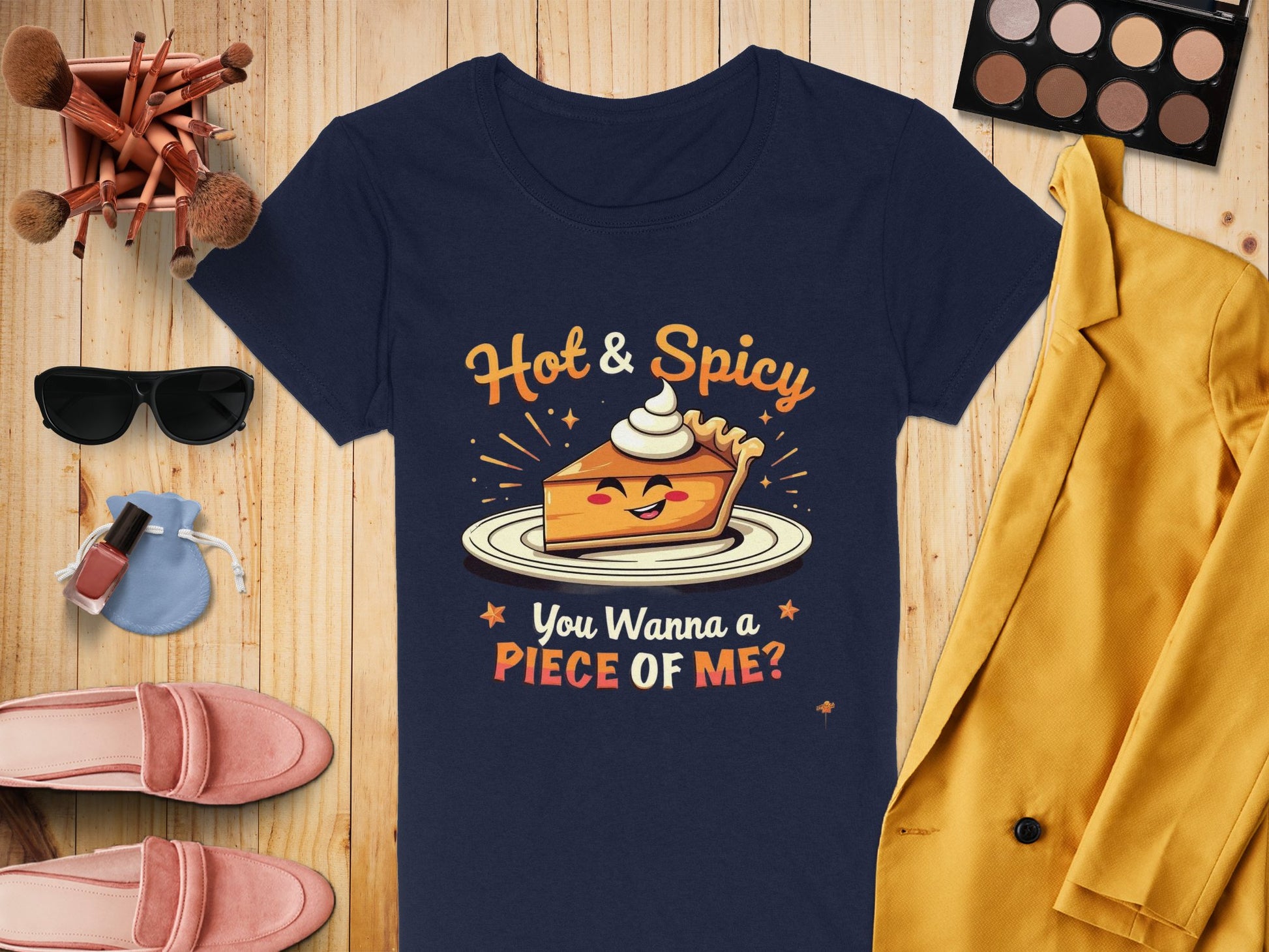 A Garment Graphics navy T-shirt showcases a pie slice with Hot & Spicy above and You Wanna a Piece of Me? below. Ideal for casual wear, its displayed with makeup, a yellow jacket, pink shoes, nail polish, and brushes on wood. Available in womens sizes.