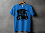 A blue T-shirt by Garment Graphics hangs against a dark gray wall, featuring a black top hat silhouette with St Patricks Day in green and black. It also includes two small green shamrocks and a subtle four-leaf clover for festive charm.