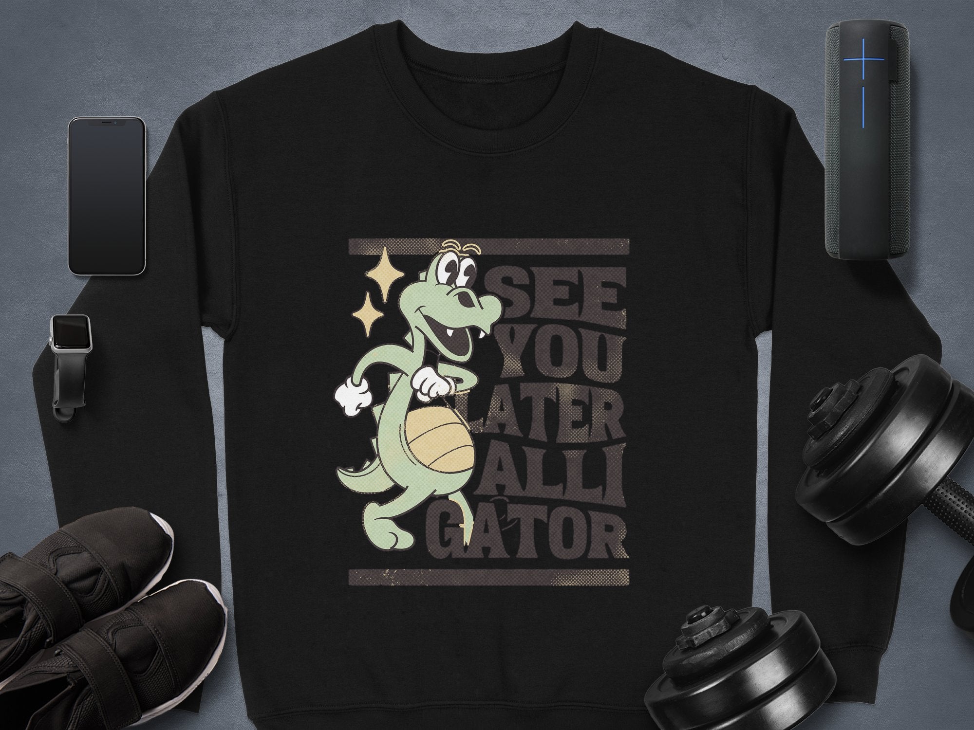 Garment Graphics black graphic sweatshirt features a cartoon alligator with a basketball, sparkle above its eye, and the text SEE YOU LATER ALLIGATOR. Unisex sizing. Styled with a phone, smartwatch, shoes, dumbbells, and speaker on a gray background.