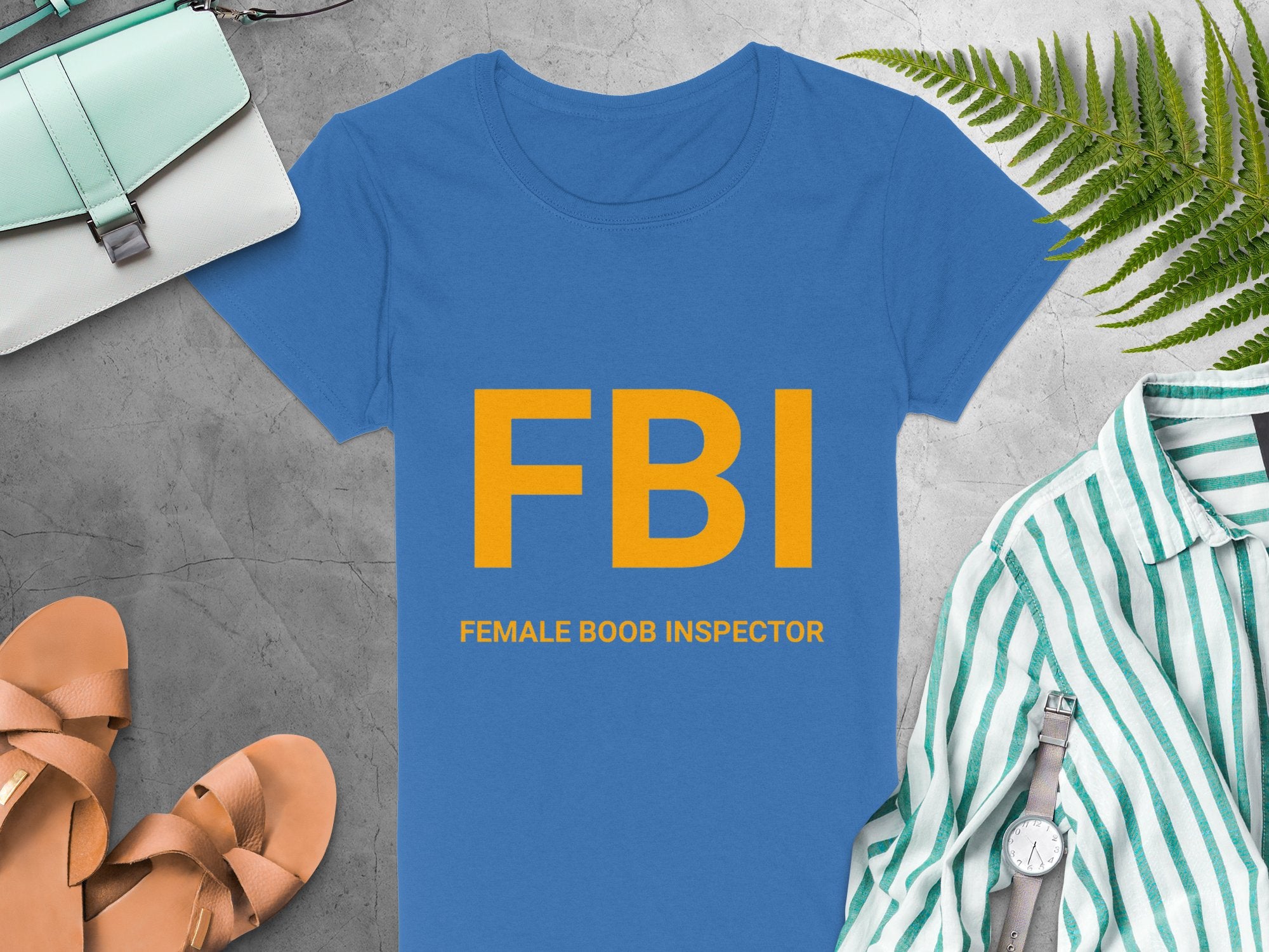 Garment Graphics blue T-shirt with FBI: Female Boob Inspector in yellow makes an ideal gag gift. Styled with a light blue purse, brown sandals, and a green and white striped shirt topped by a watch on a gray surface. Available in womens sizing for the perfect fit!.