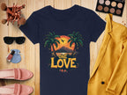 A flat lay by Garment Graphics features a navy T-shirt with a tropical sunset graphic and One Love text, paired with a yellow blazer, pink loafers, sunglasses, makeup brushes, and an eyeshadow palette—embodying chic beach vibes on a wooden surface.