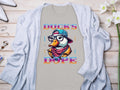 A light gray Garment Graphics T-shirt features a duck with glasses, hat, and scarf design. The text Ducks Dope is in stylized lettering. Its displayed on wood with a cardigan, flowers, and accessories nearby.