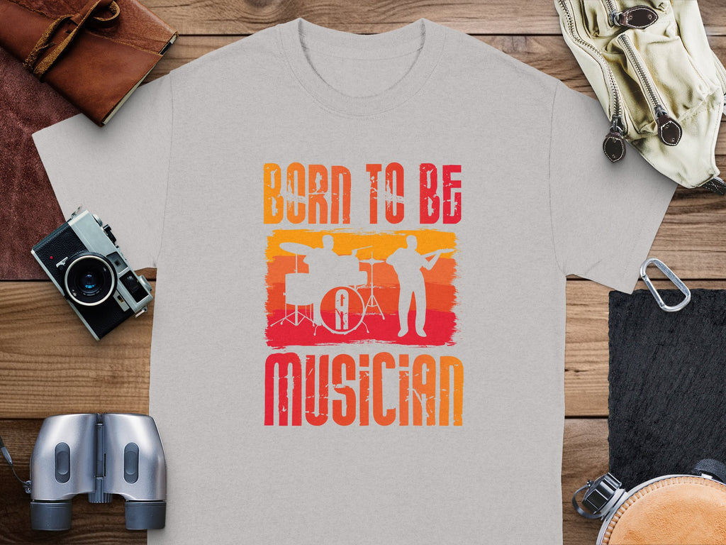 A heather grey Born to be a Musician T-shirt by Garment Graphics rests on a wooden surface, ideal for music enthusiasts. It features an orange and red graphic of a drummer and guitarist. Nearby, accessories such as a camera, leather wallets, binoculars, and coiled rope enhance the scene.