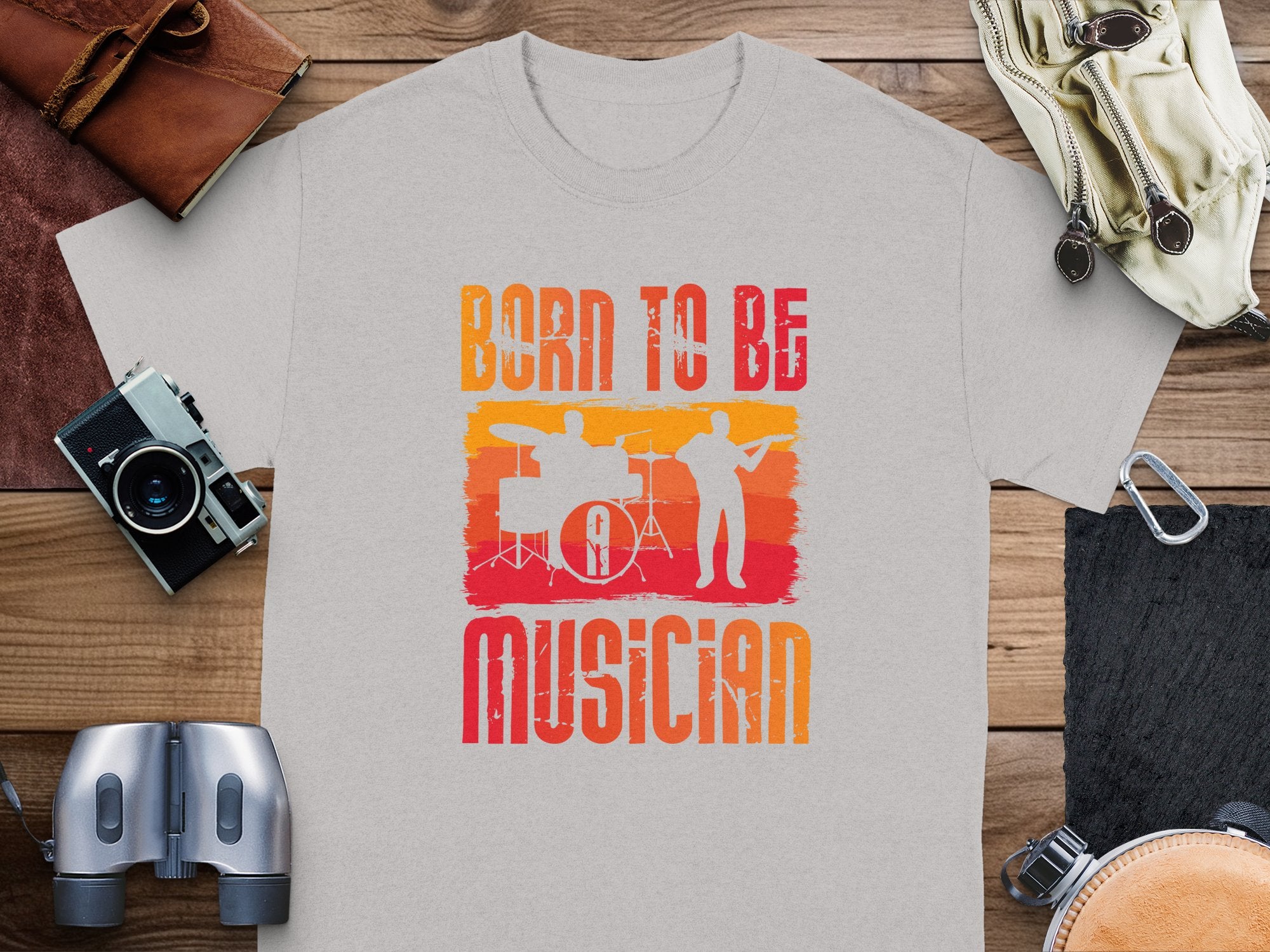A heather grey Born to be a Musician T-shirt by Garment Graphics rests on a wooden surface, ideal for music enthusiasts. It features an orange and red graphic of a drummer and guitarist. Nearby, accessories such as a camera, leather wallets, binoculars, and coiled rope enhance the scene.