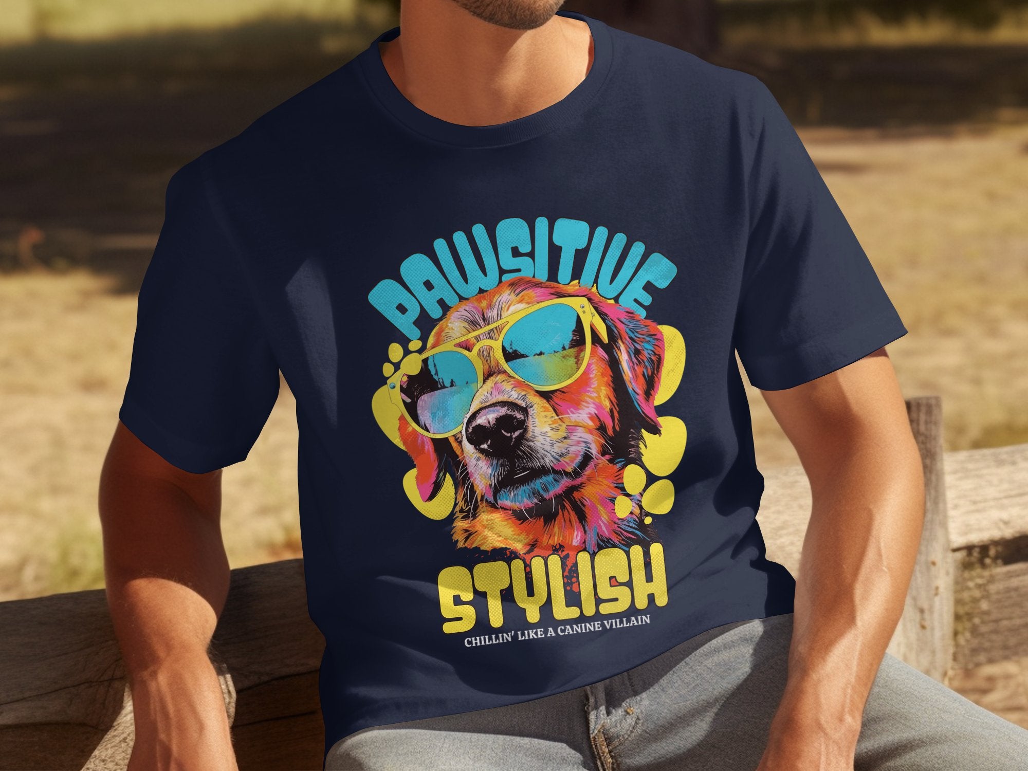 Seated on a wooden bench, a person wears a navy blue dog t-shirt by Garment Graphics featuring a colorful graphic of a dog in sunglasses. The playful phrases Paw-sitive Stylish and Chillin Like A Canine Villain are printed boldly, highlighting the essence of canine coolness.