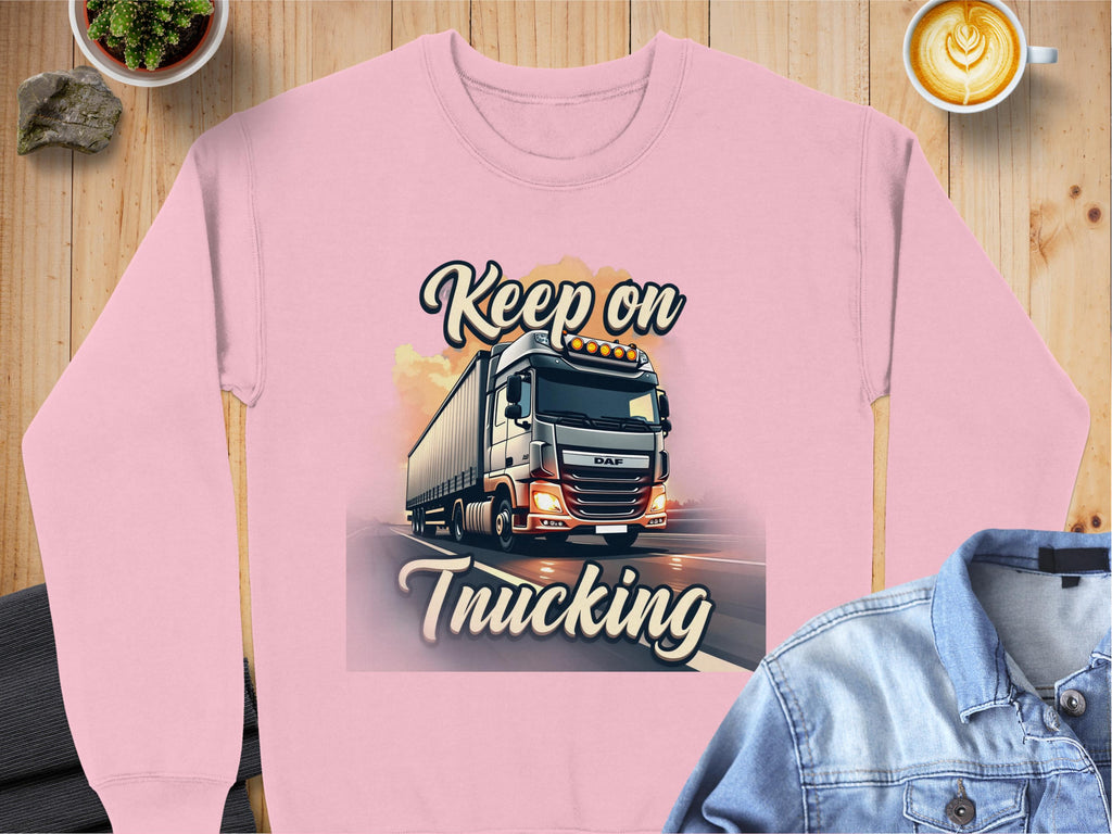 A pink Garment Graphics sweatshirt with a truck illustration and Keep on Trucking sits on a wooden surface, styled with a denim jacket and warm latte.