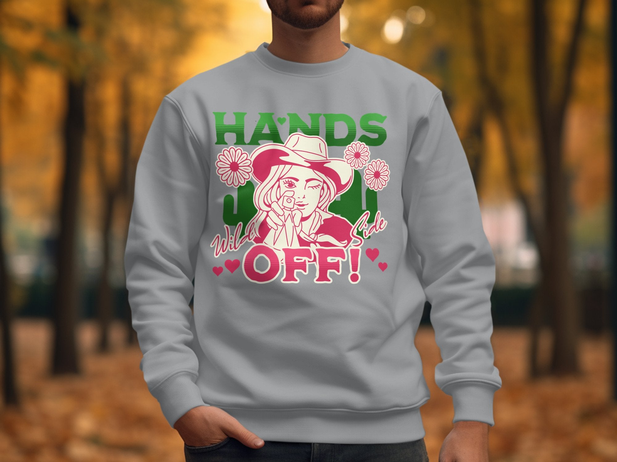 A person wears a gray sweatshirt from Garment Graphics, showcasing bold graphics of a cowboy hat and flowers. The text HANDS OFF! stands out in green and pink. Set against autumn trees with orange leaves, this unisex piece is perfect for any wardrobe.